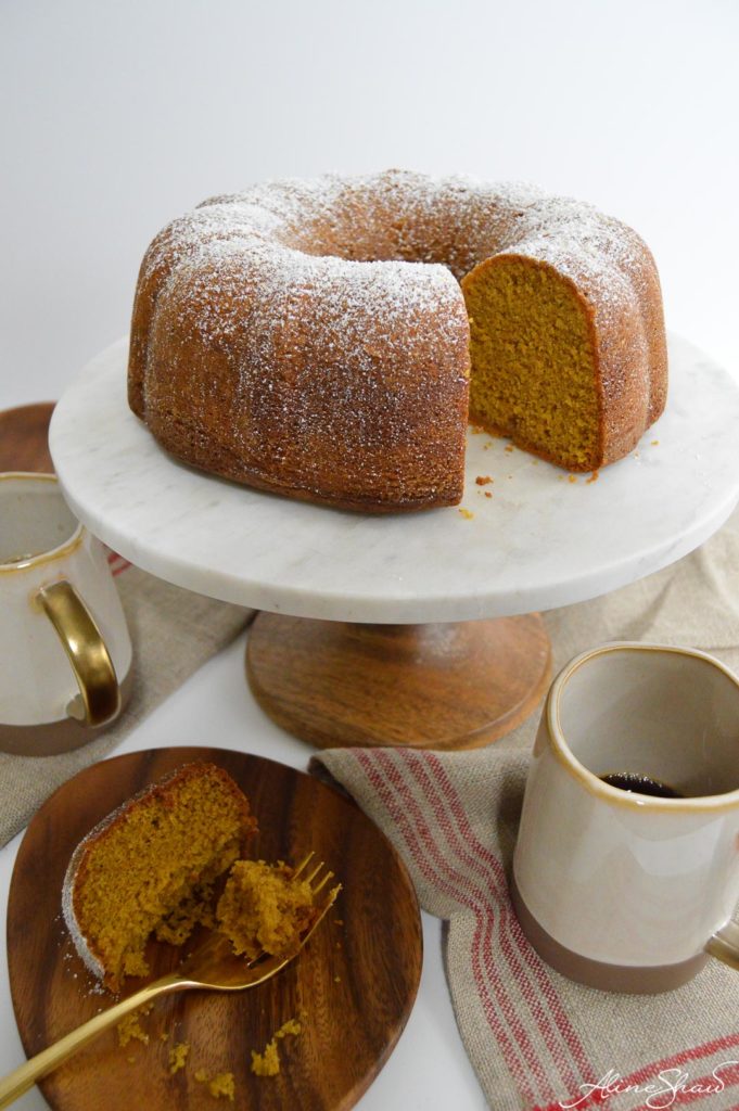 A bundt cake on a white cake stand next to a plate of cake and a cup of coffee