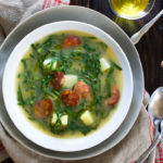 A bowl of Portuguese green soup in a white and grey bowl next to two spoons on a tabletop