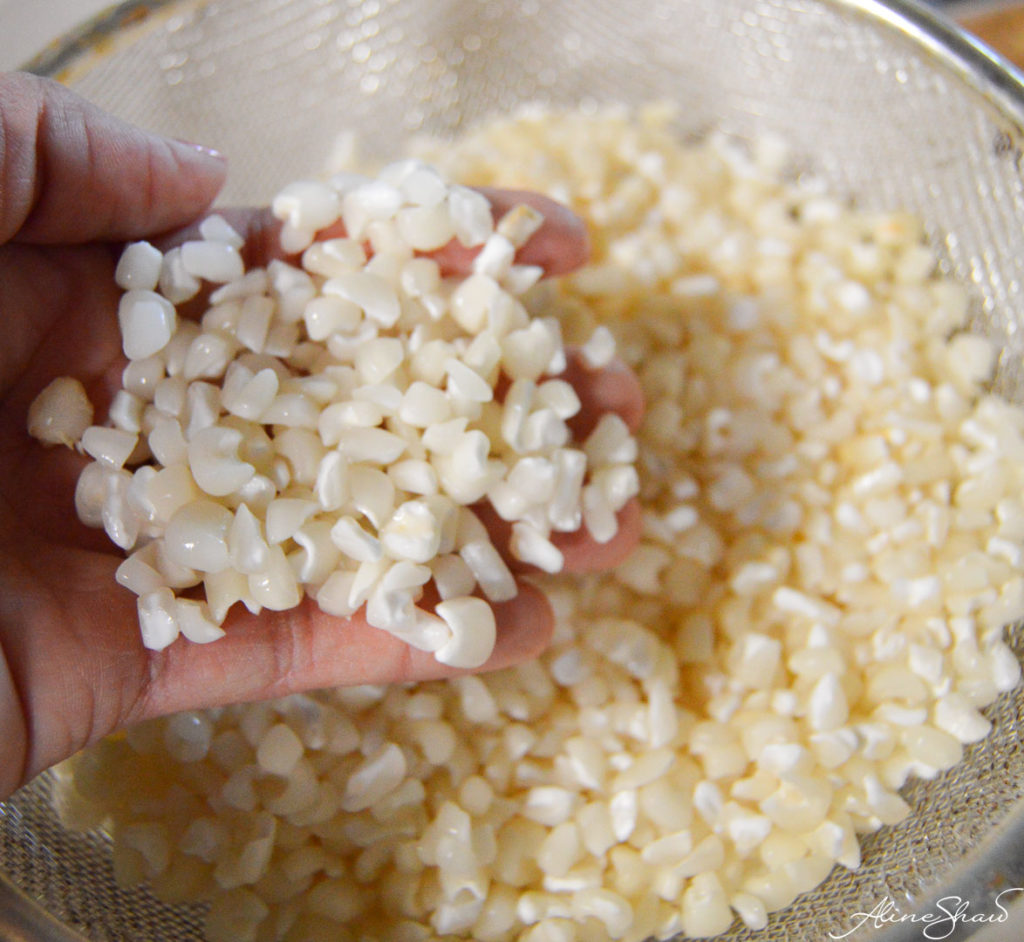 A hand holds rinsed white hominy corn above a fine mesh strainer