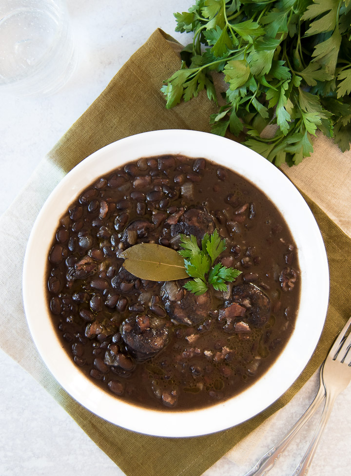 A white bowl holds Brazilian Black Beans, garnished with parsley and a bay leaf