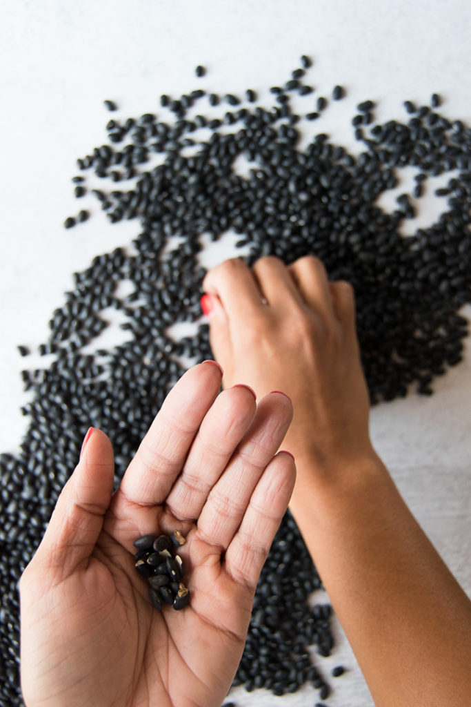 A woman holds broken black beans in the palm of her hand above more scattered beans on a kitchen surface