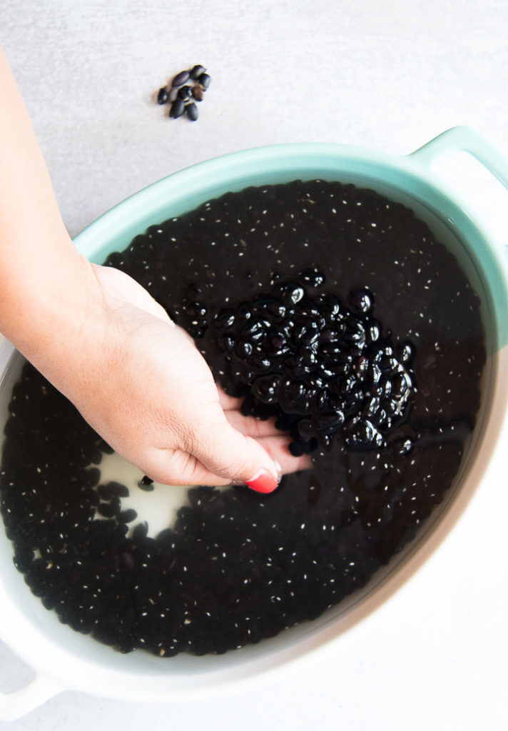 A woman scoops her hands into a turquoise container where black beans are soaking