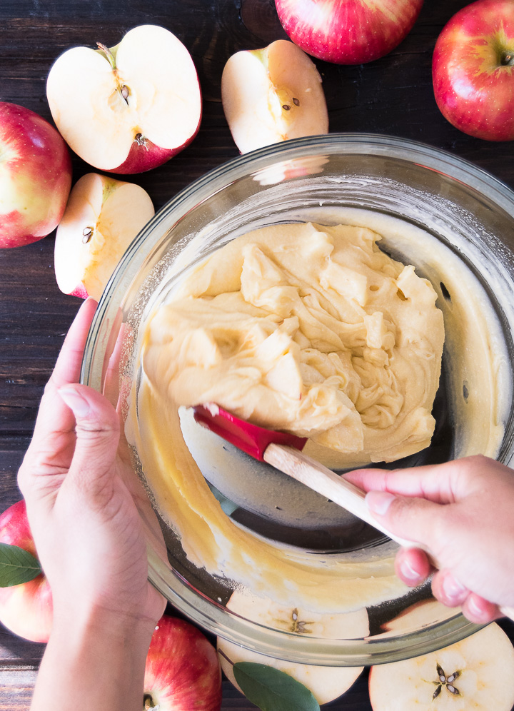 A whisk folds apple chunks into cake batter in a bowl