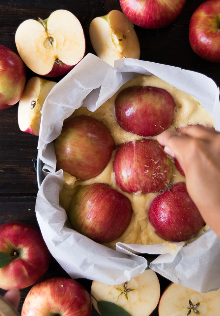 Halved apples on top of cake batter in a round pan are sprinkled with a sugar mixture