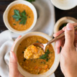 a set of hands holds a spoonful of lentil soup above a white bowl