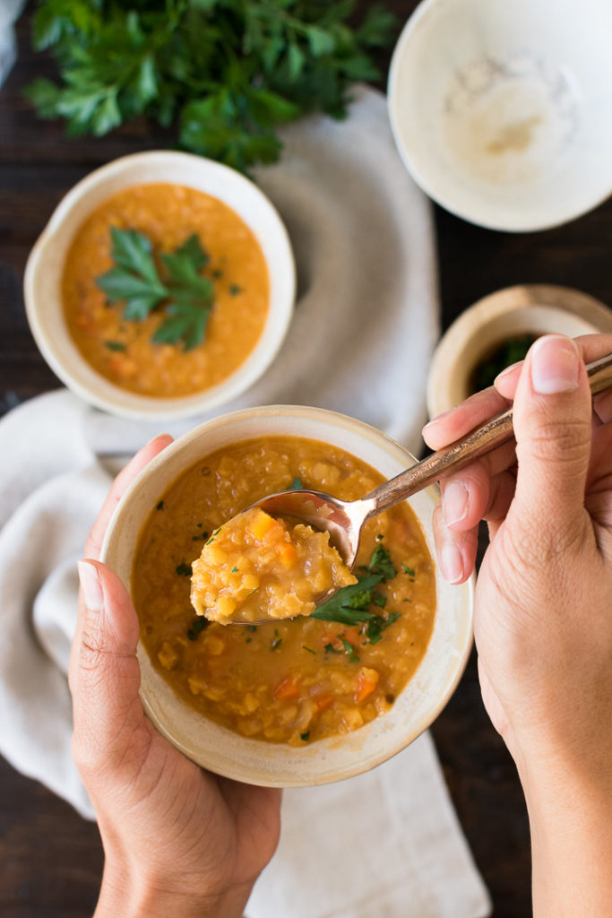 a set of hands holds a spoonful of lentil soup above a white bowl 