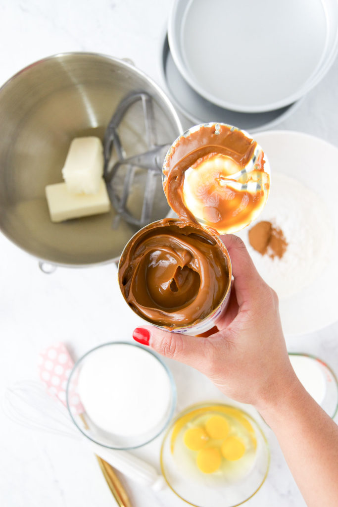 A hand holds an open can of La Lechera dulce de leche above cake ingredients in bowls