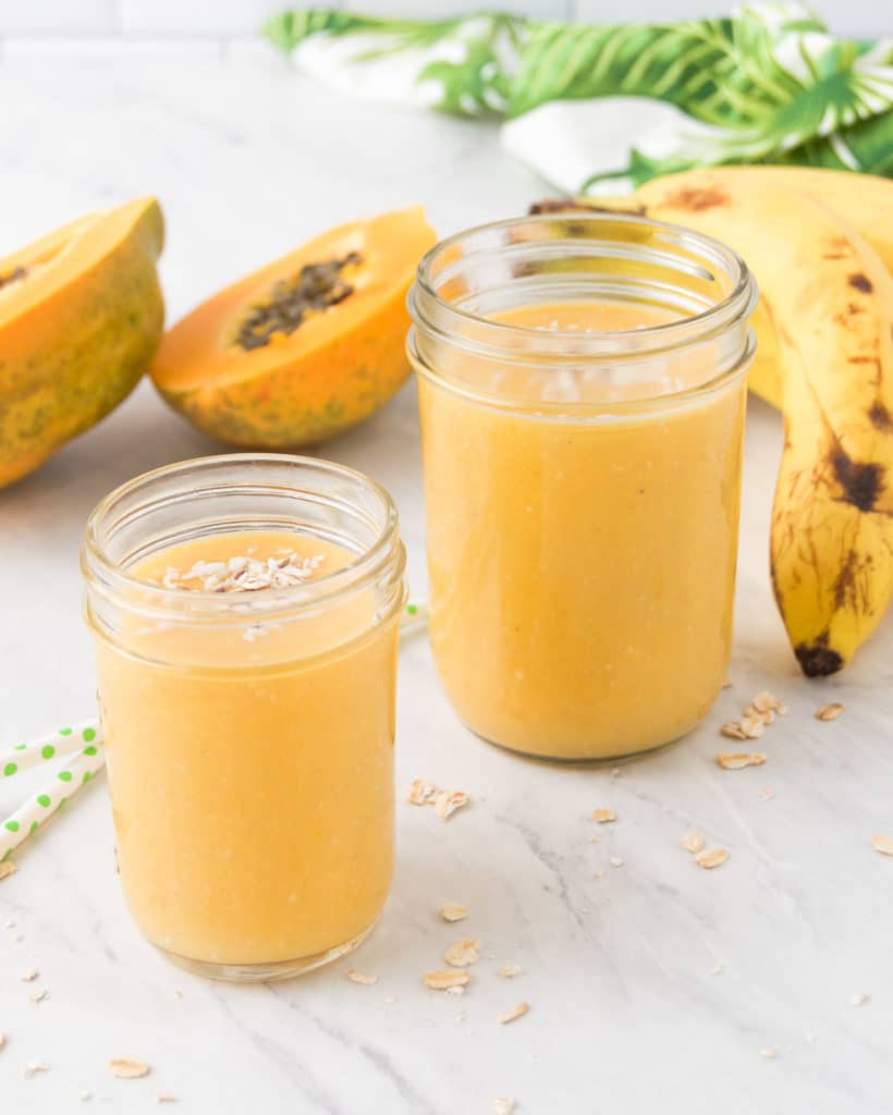 two mason jars holding tropical smoothies on marble, surrounded by banana and papaya
