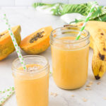 two papaya banana smoothies in mason jars on a marble countertop surrounded by fresh fruit