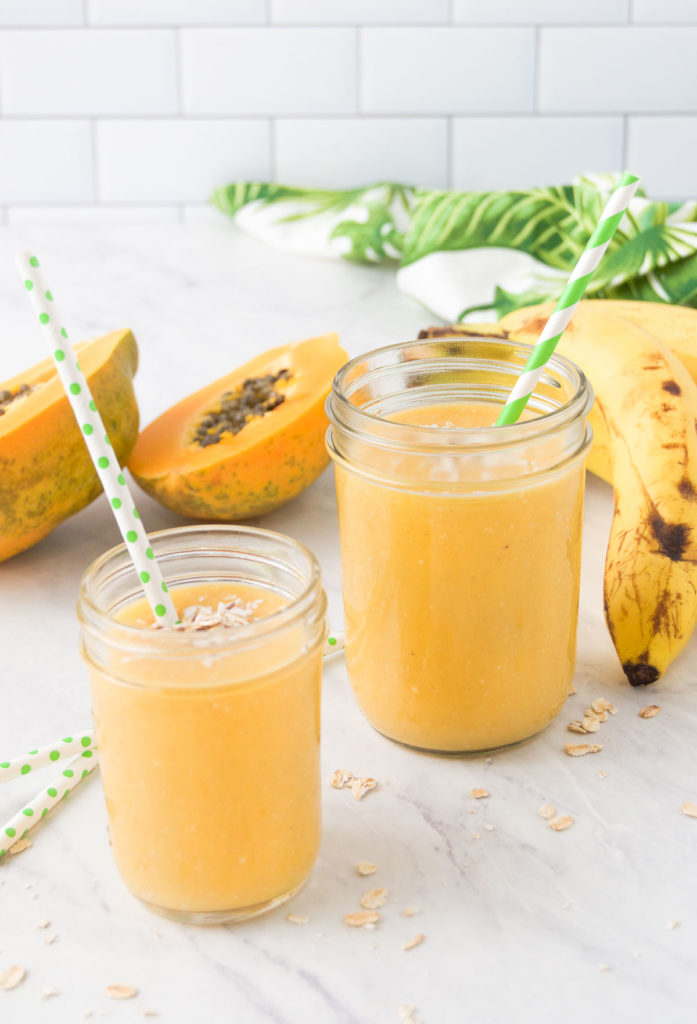 two papaya banana smoothies in mason jars on a marble countertop surrounded by fresh fruit
