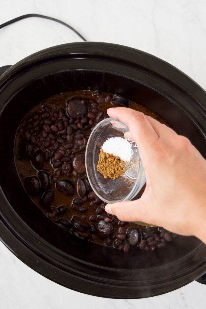 A woman pours cumin, salt and pepper into a slow cooker full of black beans