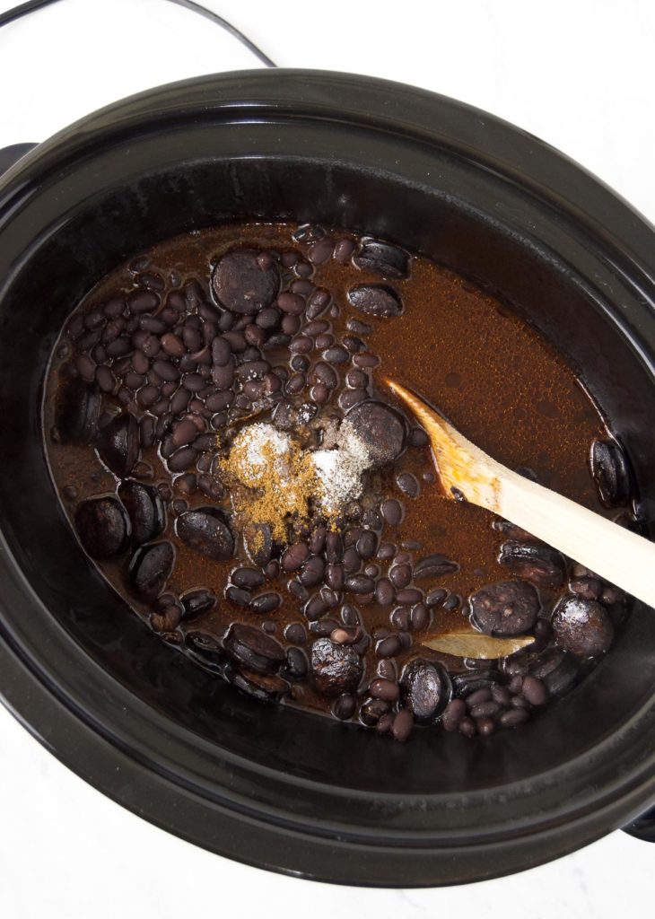 A wooden spoon stirs seasonings into crockpot black beans