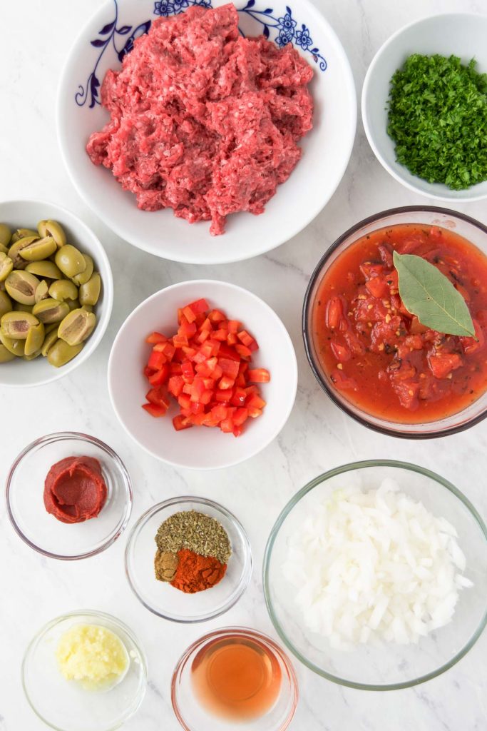 Ground beef, green olives, bell pepper, onion and other picadillo recipe ingredients in bowls from above