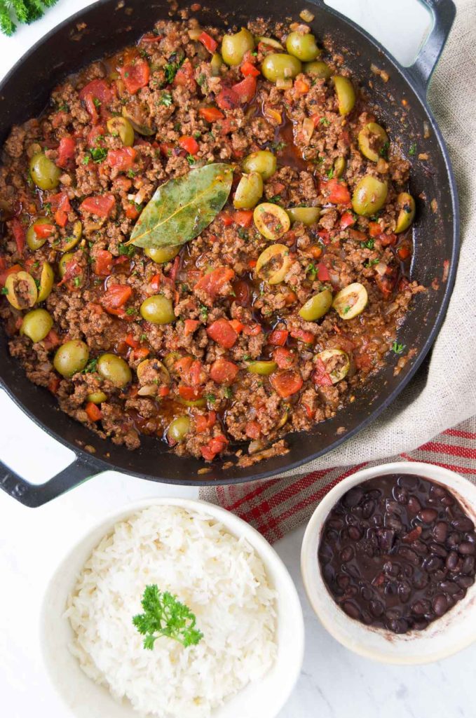 Brazilian picadillo in a black braising pan sits next to a white bowl of rice and a small white bowl holding black beans