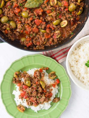 Picadillo is served over rice in a bright green bowl next to a pan holding more picadillo and a bowl holding extra rice