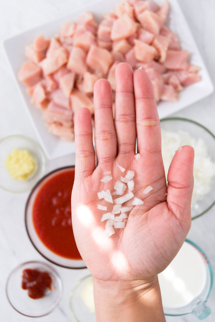 A hand holds diced onions above ingredients for a chicken recipe