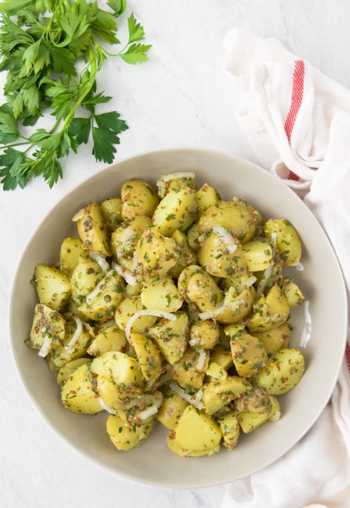 A white bowl holds vegan potato salad next to a bunch of parsley