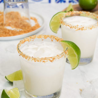 Two Coconut Margaritas garnished with coconut and lime on marble next to colorful blue tile