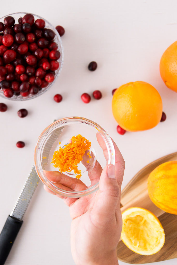 A woman holds a bowl of orange zest above a bowl of cranberries
