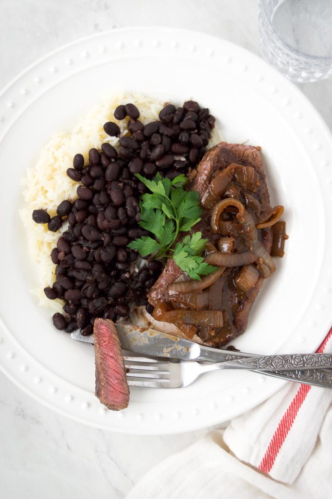 A fork holds a slice of steak next to the whole steak with caramelized onions, and a pile of beans and rice