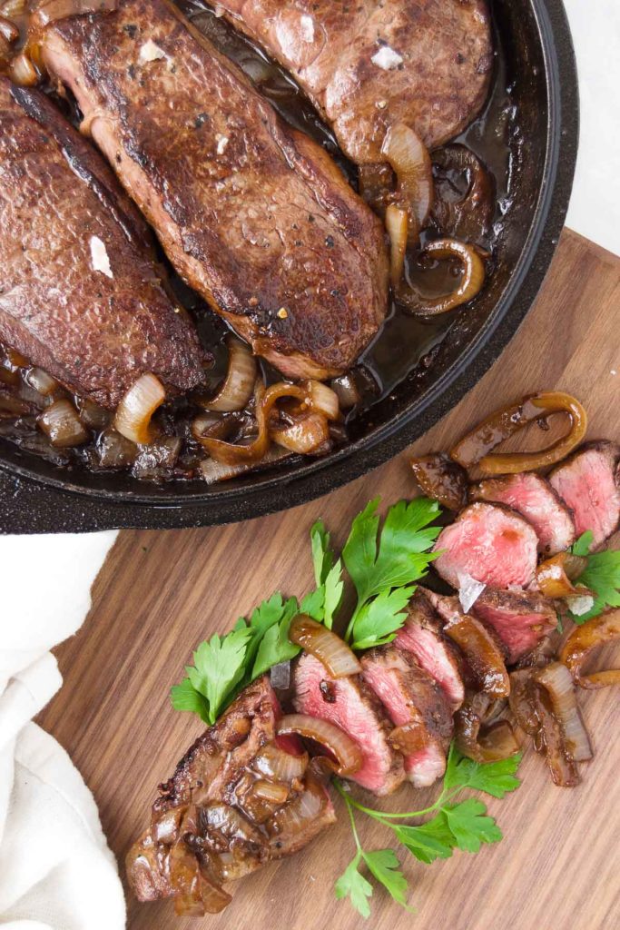 Cooked steaks in a pan with caramelized onions next to a cutting board with sliced steak, herbs and onions
