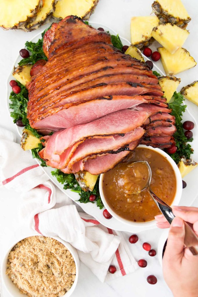 A person lifts a spoon of pineapple ham glaze sitting next to a roasted ham