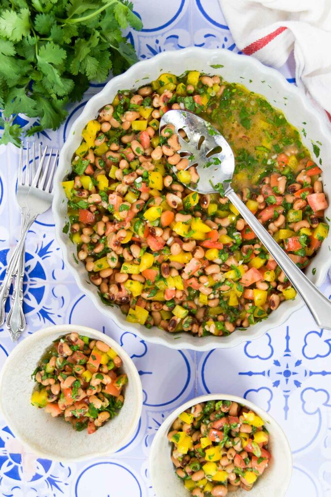 A bowl of black eyed peas salad on a tile background with a spoon, surrounded by herbs and two other small serving bowls