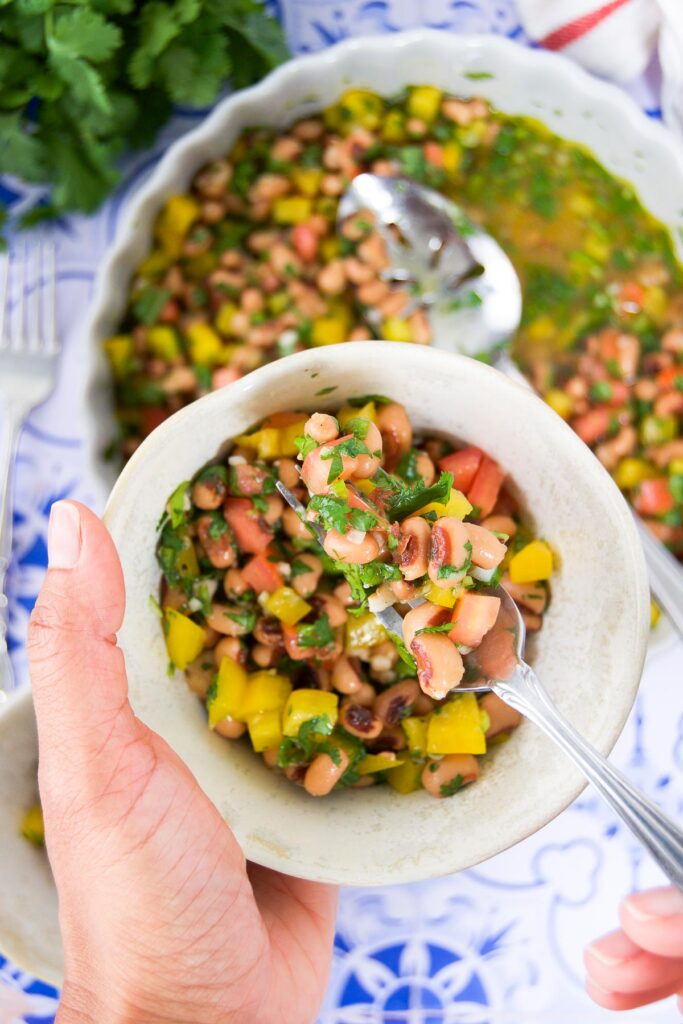 A woman holds a serving bowl of black eyed pea salad along with a fork