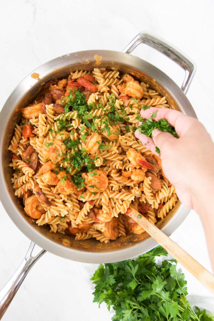 A woman sprinkles fresh parsley on top of a pan of sausage and shrimp pasta