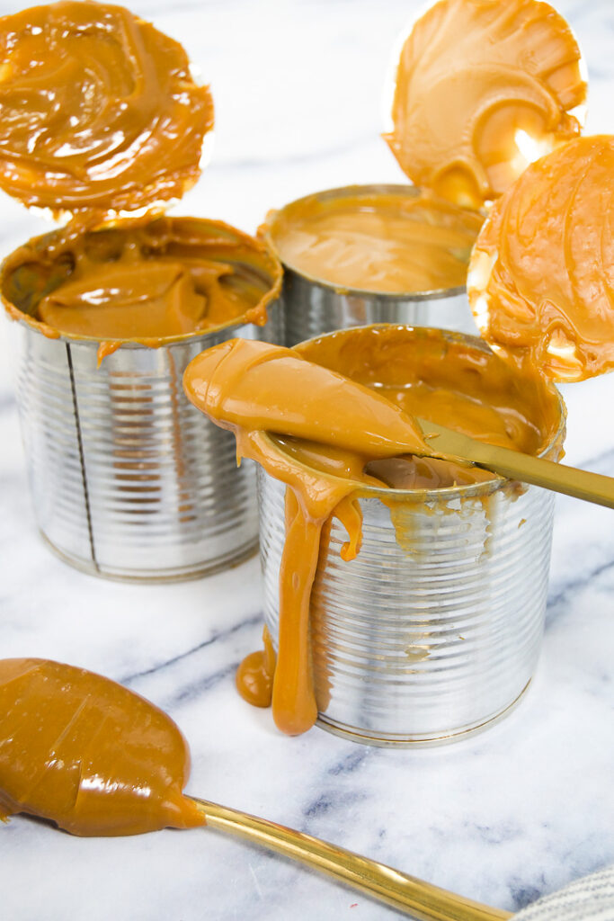 A butter knife with dulce de leche on it on top of an open can with two other cans of dulce de leche on marble
