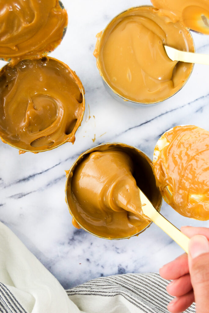 A spoon stirs a can of dulce de leche as it sits on the counter next to two other opened cans