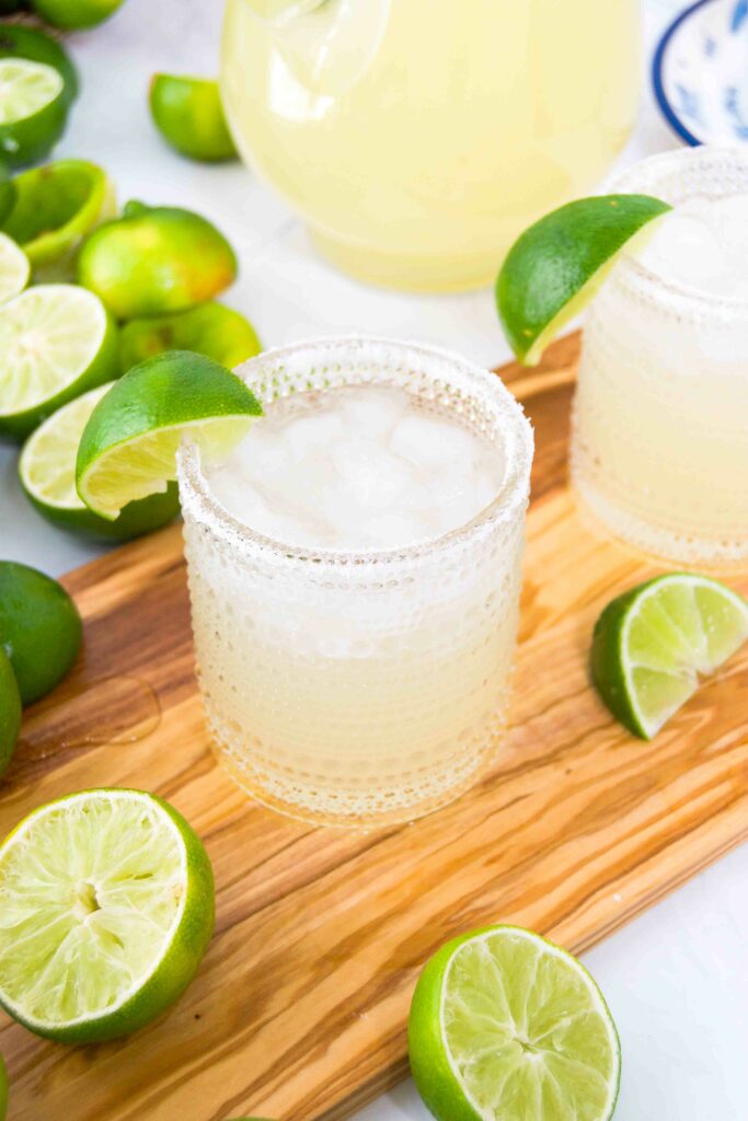 Two margaritas on a wooden cutting board surrounded by sliced limes