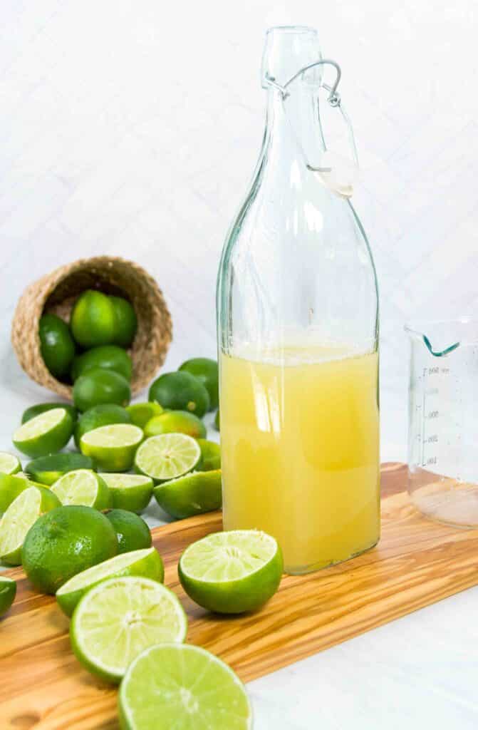 A container of margarita mix on a wooden cutting board surrounded by halved limes