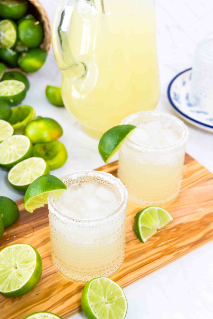 Two glasses of margaritas garnished with lime wedges and salt sit on a wooden cutting board with lots of halved limes and a pitcher