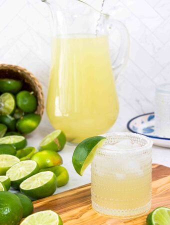 A single glass of margarita in front of a pitcher of margaritas on a wooden cutting board with limes