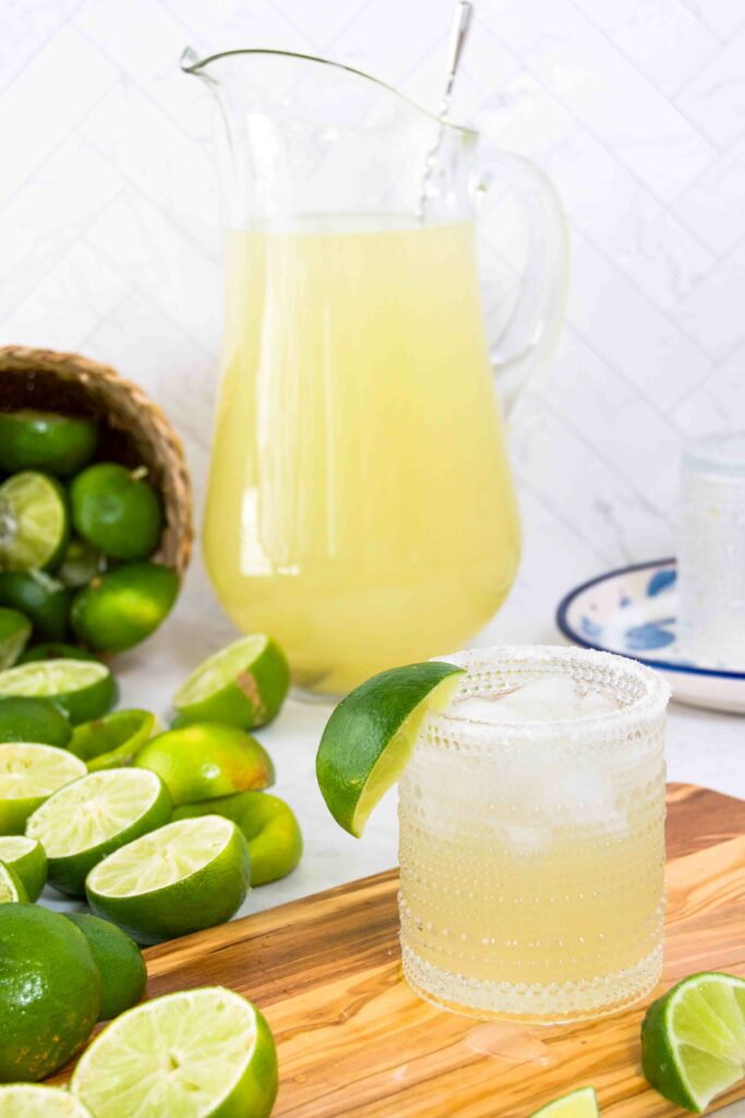 A single glass of margarita in front of a pitcher of margaritas on a wooden cutting board with limes