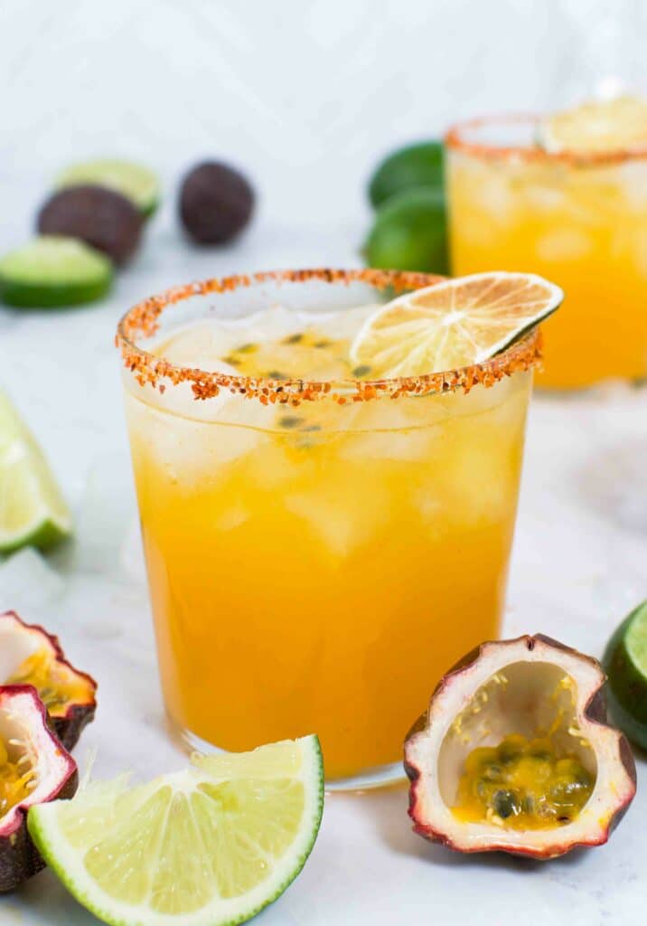Close up of a passion fruit margarita rimmed with tajin with a dried lime on top on marble surrounded by limes and passion fruits