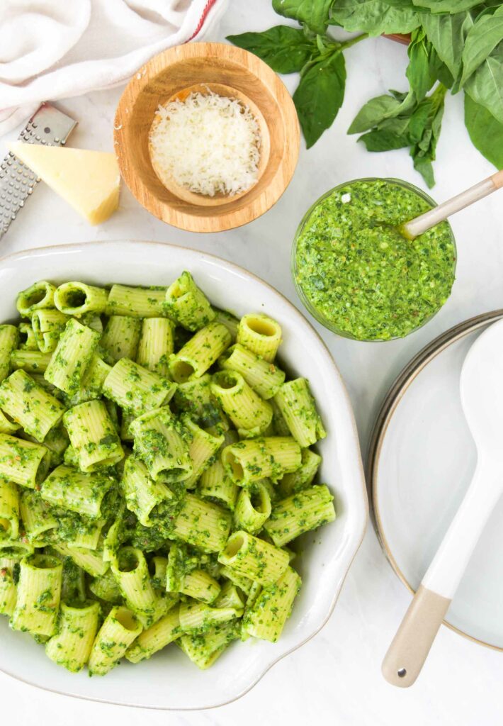 A large bowl of basil pesto pasta next to smaller bowls of grated cheese and pesto sauce