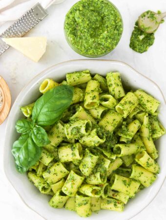 A bowl of pesto pasta garnished with fresh basil leaves, next to bowls of pesto, grated parmesan and a block of cheese