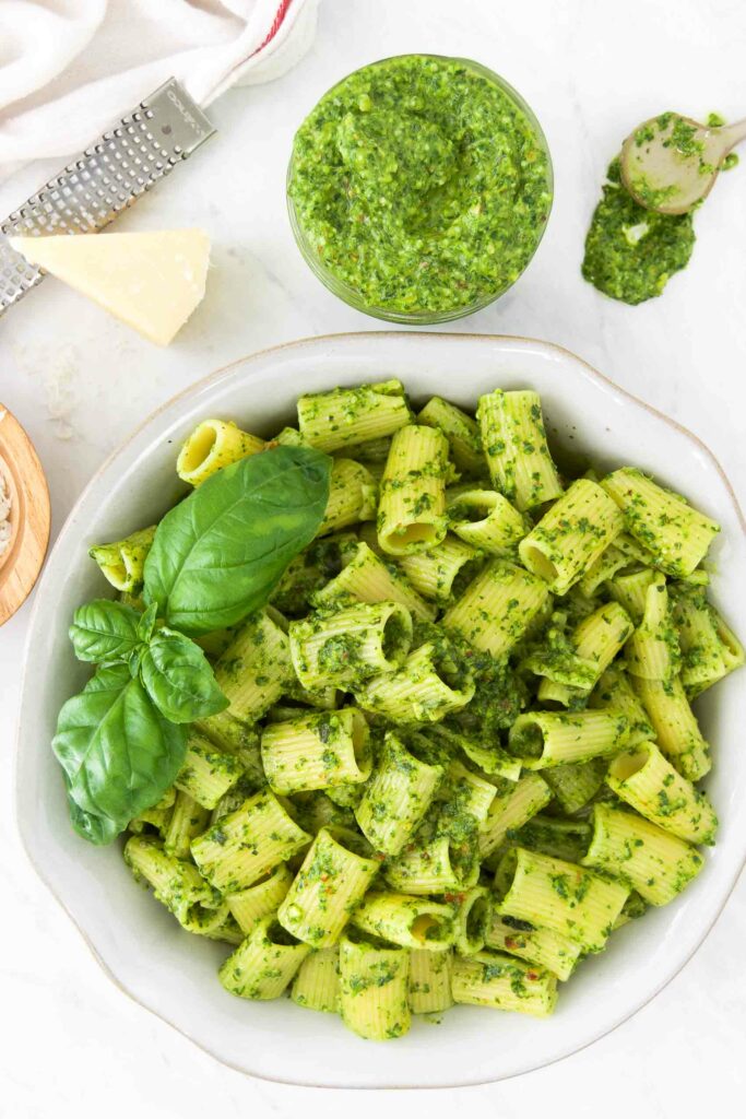 A bowl of pesto pasta garnished with fresh basil leaves, next to bowls of pesto, grated parmesan and a block of cheese