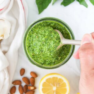 A hand lifts a spoonful of spinach pesto out of a jar on a white surface surrounded by lemon, almonds, greens and a linen