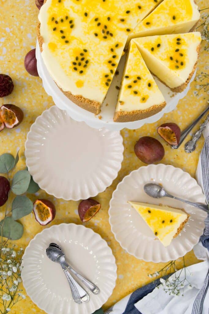 overhead of a sliced passion fruit cheesecake on a stand next to scalloped plates with slices and forks