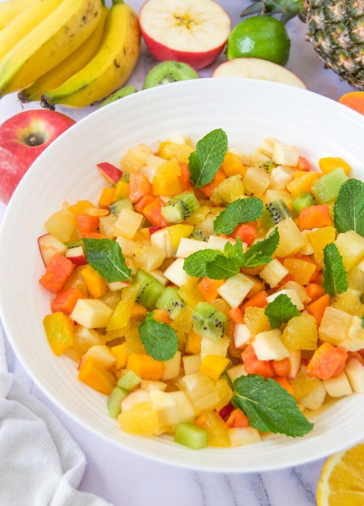 a white bowl of an easy fruit salad with tropical fruits garnished with mint leaves and surrounded by more fruits