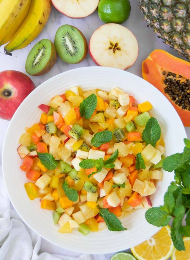 a white bowl holding diced tropical fruit salad surrounded by halved tropical fruits on marble