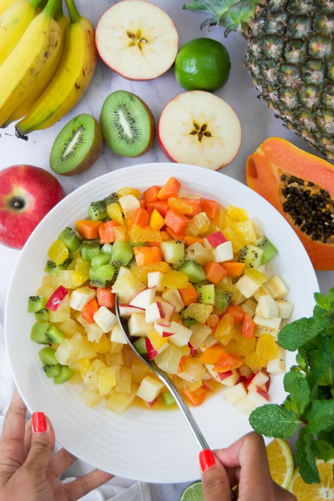 a spoon stirs an easy fruit salad surrounded by halved tropical fruits