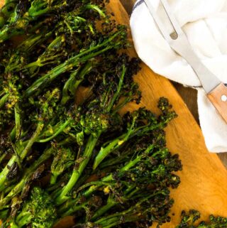 charred grilled broccolini on a wooden cutting board next to grilling tools and a linen