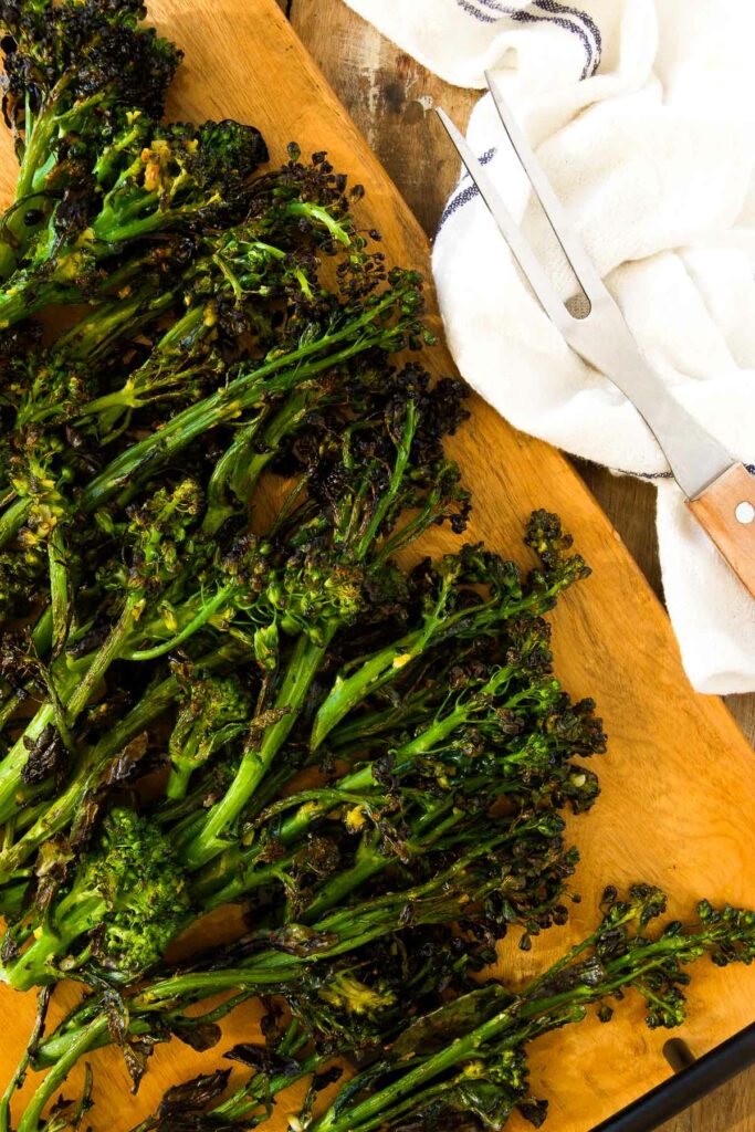 charred grilled broccolini on a wooden cutting board next to grilling tools and a linen