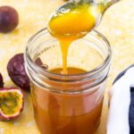 A spoon drizzles passion fruit sauce into a mason jar sitting on a yellow surface surrounded by halved purple passion fruits