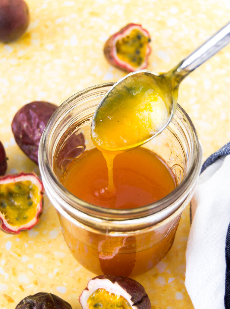 Passion fruit sauce drizzles from a spoon into a mason jar on a yellow surface