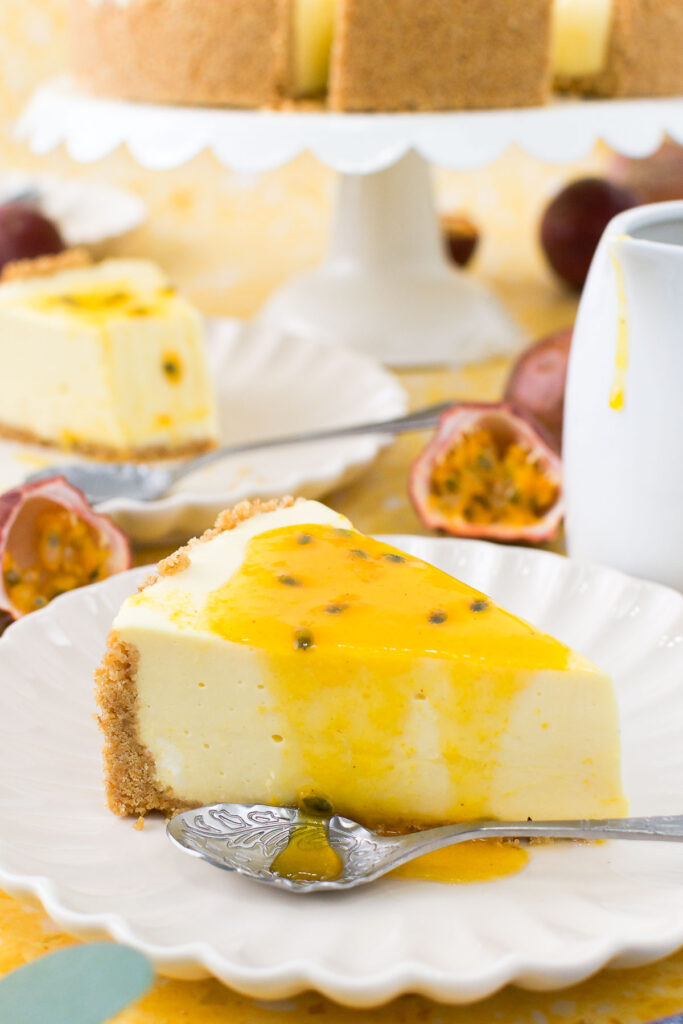 close up of a slice of passion fruit cheesecake on a scalloped plate with a fork, topped with fresh passion fruit, on a yellow terrazzo surface